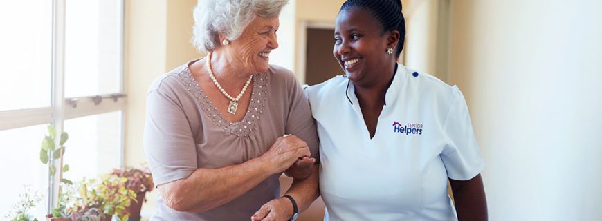 Smiling home caregiver and senior woman walking together Portrait of smiling home caregiver and senior woman walking together through a corridor. Healthcare worker taking care of elderly woman.